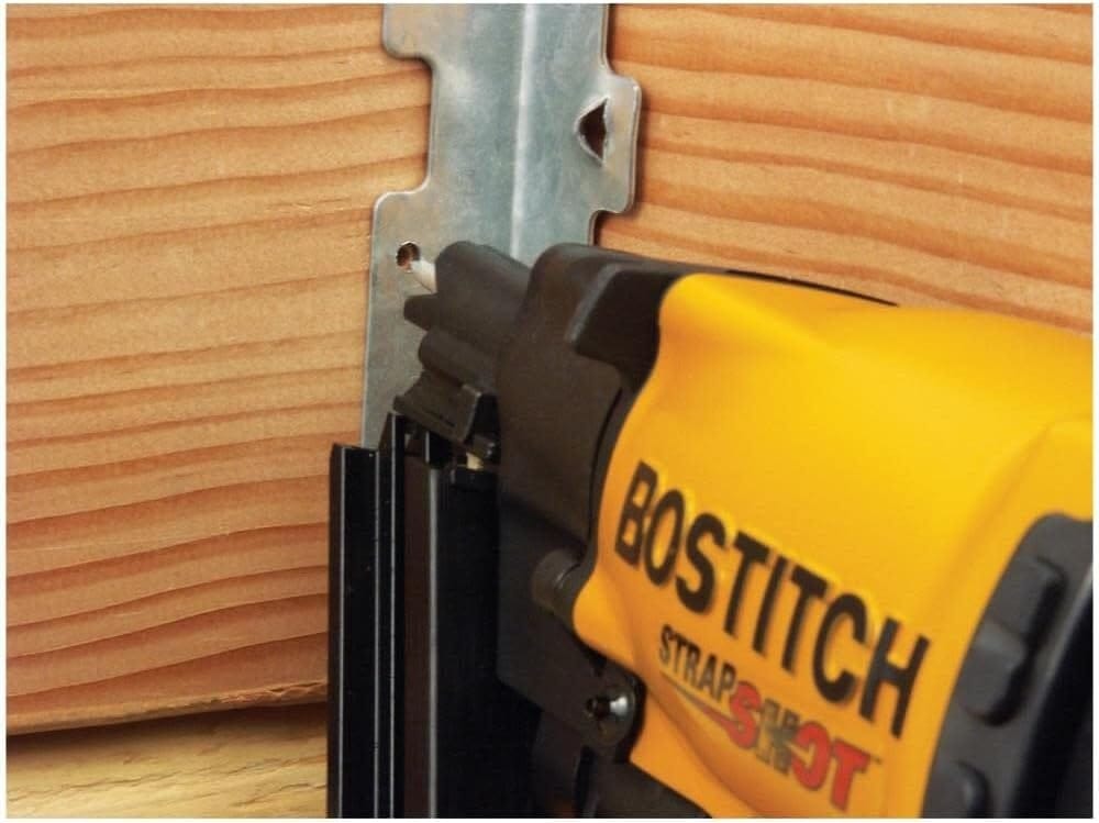 Action close-up showing the Bostitch MCN150 pneumatic nailer fastening a metal hurricane tie to a wooden framing member, emphasizing the compact size and maneuverability in tight structural spaces.