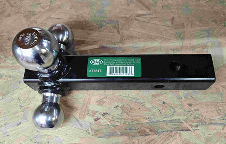 Close-up of a Brophy triple ball mount showing three chrome hitch balls on a black metal base. The center ball is engraved with “BROPHY 2 5/16" 10000 LBS,” indicating size and towing capacity. A green label with barcode is partially visible. The mount rests on a textured wooden surface.
