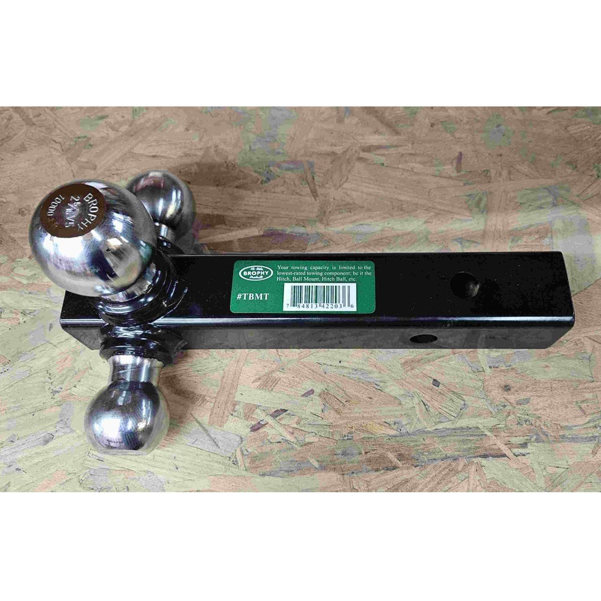 Close-up of a Brophy triple ball mount showing three chrome hitch balls on a black metal base. The center ball is engraved with “BROPHY 2 5/16" 10000 LBS,” indicating size and towing capacity. A green label with barcode is partially visible. The mount rests on a textured wooden surface.