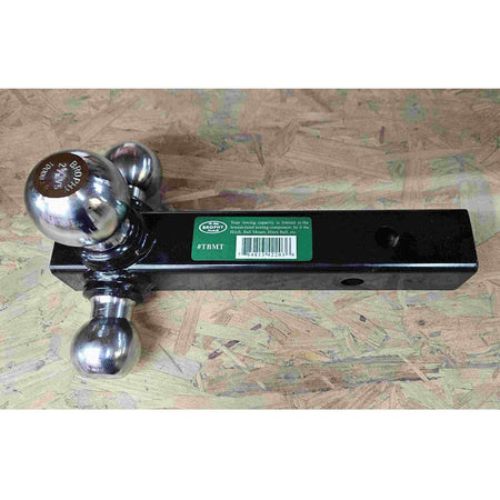Close-up of a Brophy triple ball mount showing three chrome hitch balls on a black metal base. The center ball is engraved with “BROPHY 2 5/16" 10000 LBS,” indicating size and towing capacity. A green label with barcode is partially visible. The mount rests on a textured wooden surface.