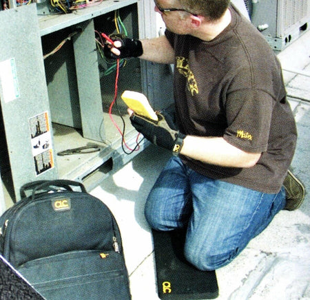 An HVAC technician kneeling on the CLC kneeling pad while wearing gloves and using a multimeter to check the wiring on an electrical unit.