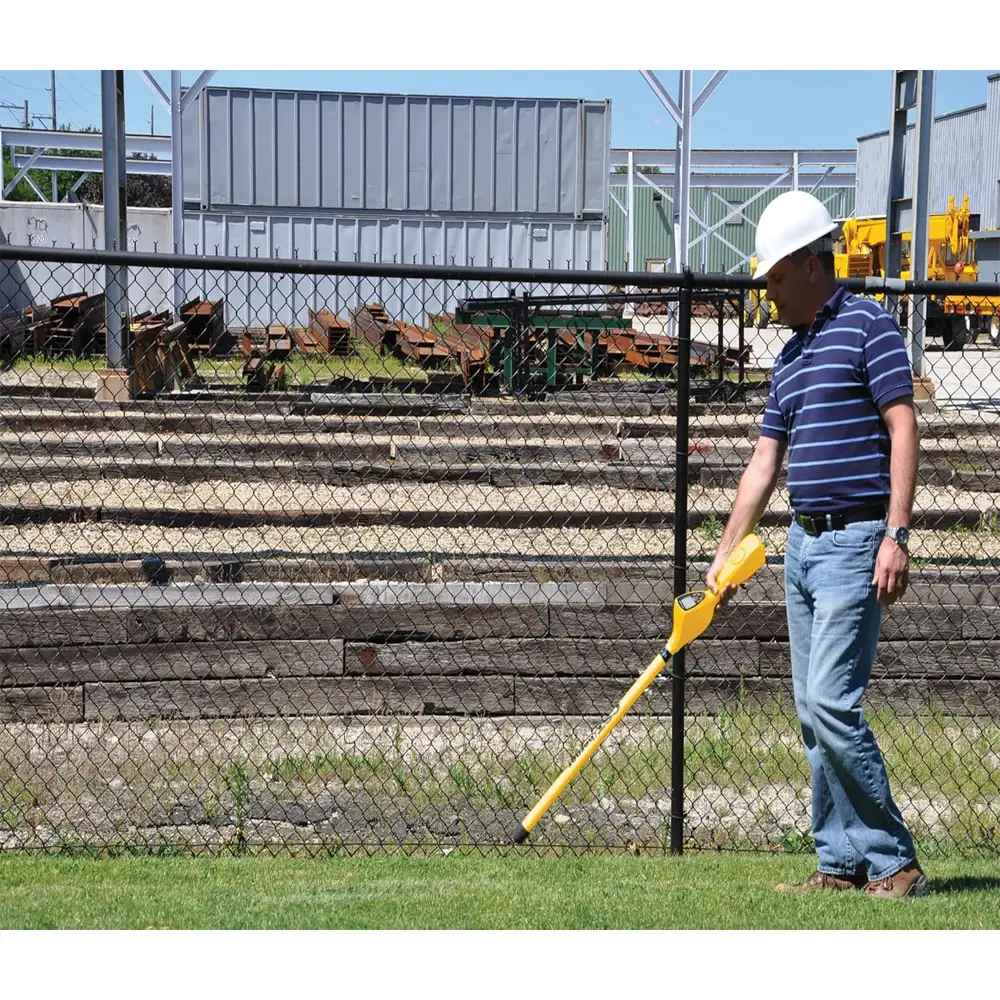 Person wearing a white hard hat using a yellow CST/berger Magna-Trak 200 magnetic locator on grass near a chain-link fence and industrial buildings.