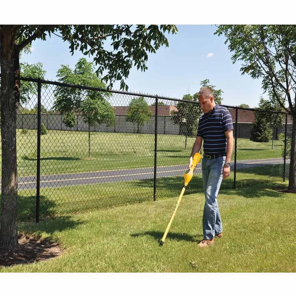 Individual in striped shirt and jeans scanning grassy ground with a yellow CST/berger Magna-Trak 200 magnetic locator near a black chain-link fence.