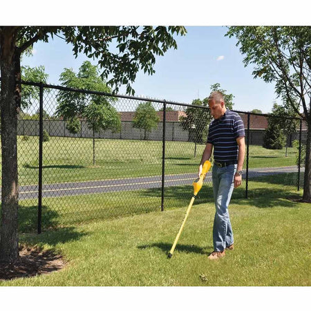 Individual in striped shirt and jeans scanning grassy ground with a yellow CST/berger Magna-Trak 200 magnetic locator near a black chain-link fence.