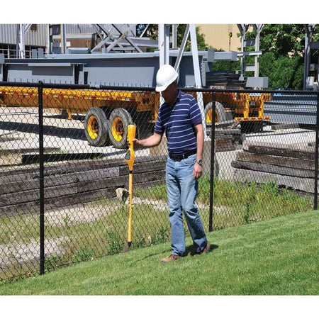 Worker in hard hat and striped polo shirt using a yellow CST/berger Magna-Trak 200 magnetic locator beside a fence with industrial equipment in the background.
