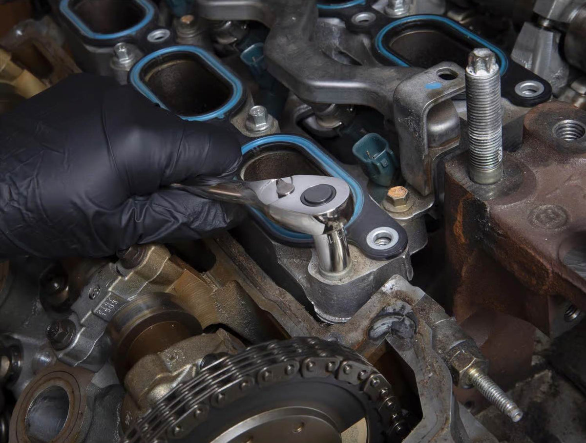 A close-up of a person wearing black gloves using a chrome ratchet and socket to tighten a bolt on a car engine, showing intricate engine components.