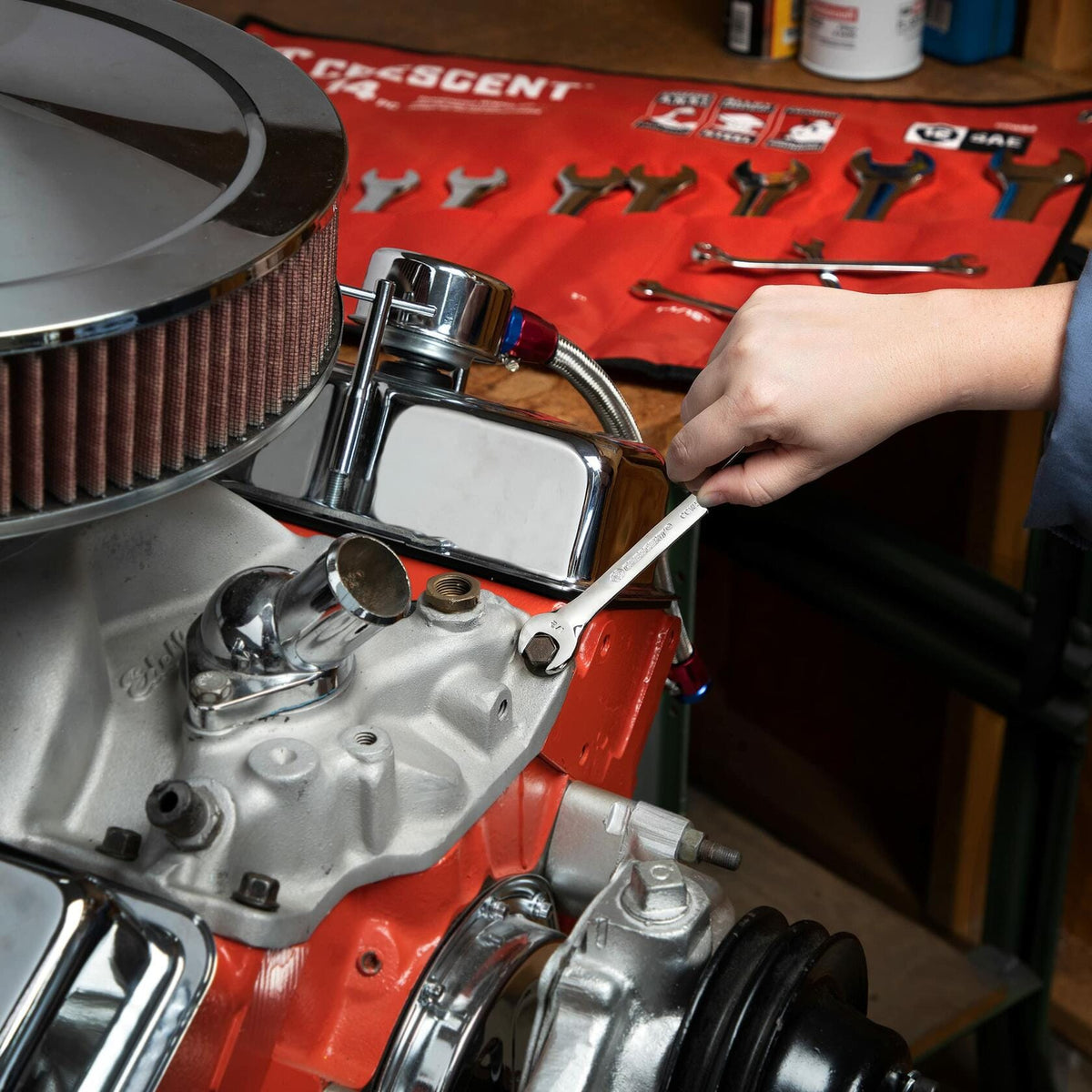 Person using a Crescent wrench to adjust a bolt on a V8 engine with a chrome air filter and red block. A red Crescent tool roll with organized wrenches is visible in the background.