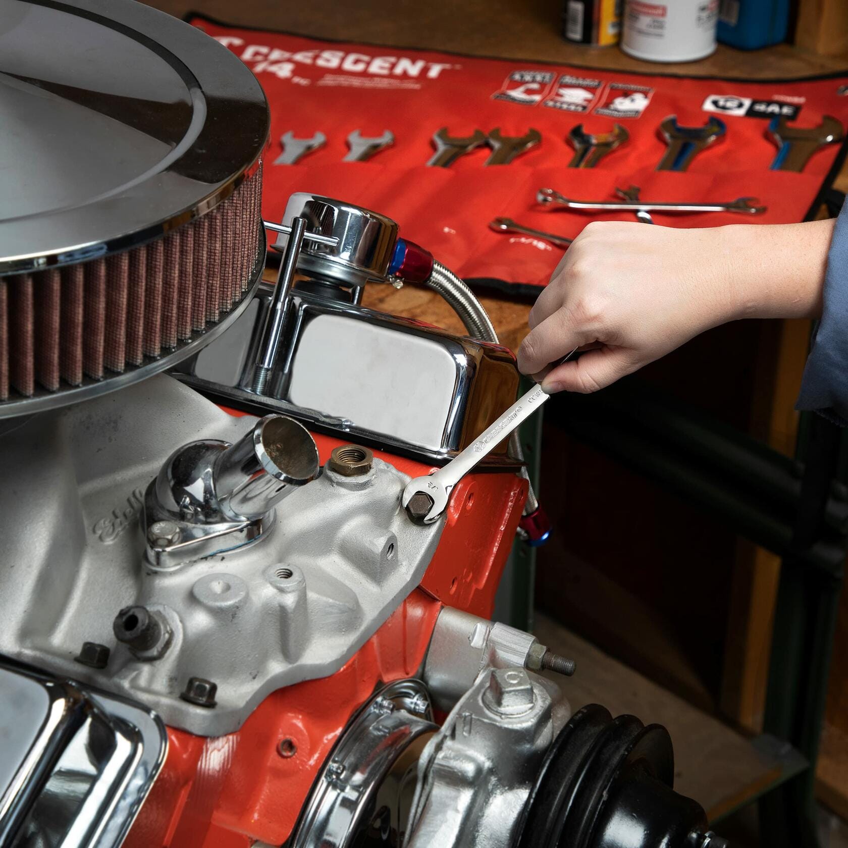 Person using a Crescent wrench to adjust a bolt on a V8 engine with a chrome air filter and red block. A red Crescent tool roll with organized wrenches is visible in the background.