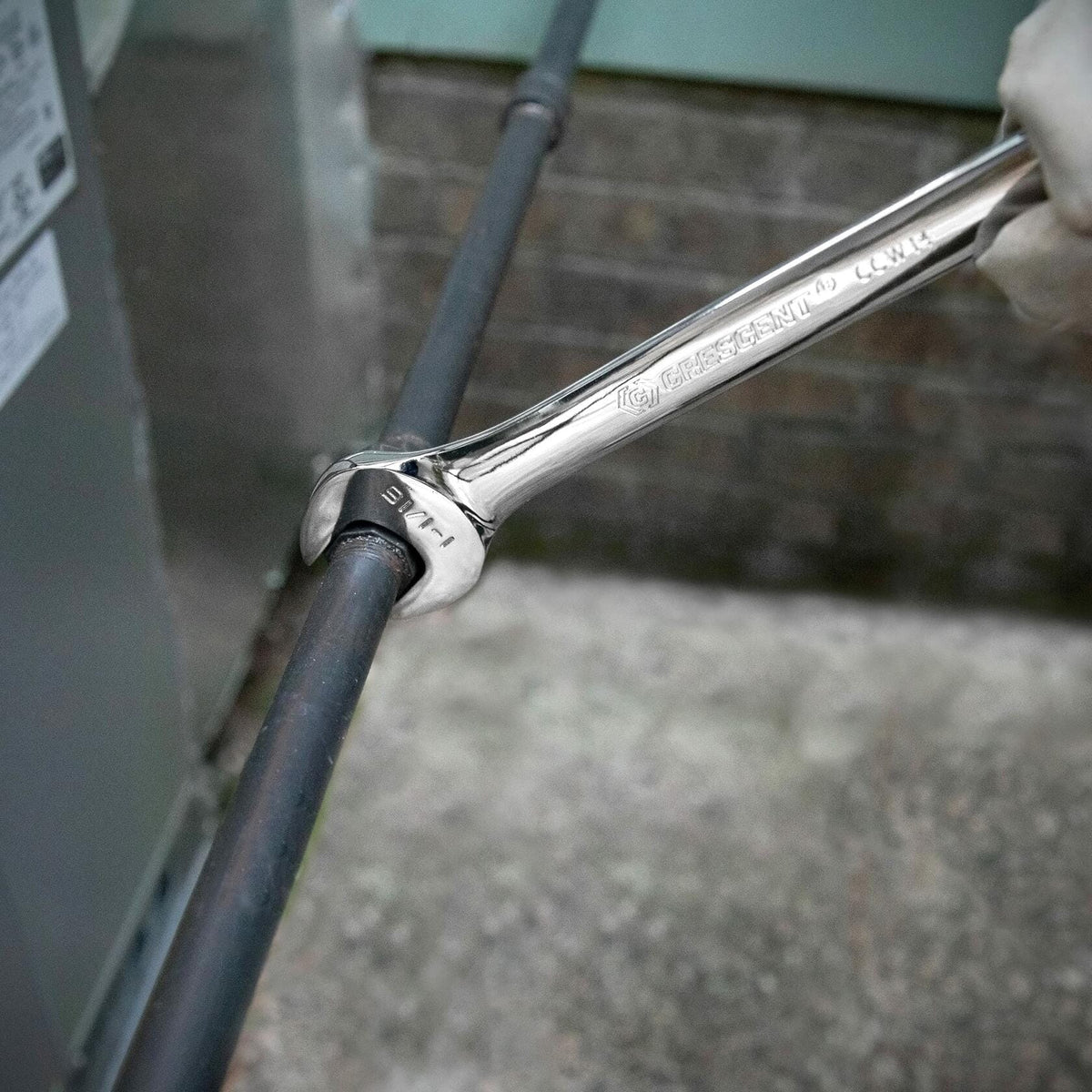 Close-up of a Crescent 7/8" combination wrench being used to adjust a pipe fitting. The background includes a brick wall and metal appliance, suggesting HVAC or plumbing maintenance.