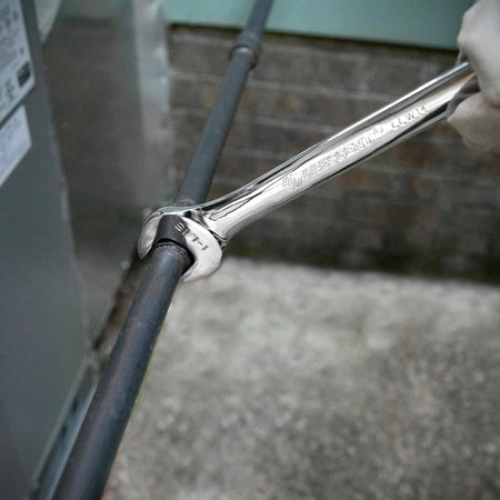 Close-up of a Crescent 7/8" combination wrench being used to adjust a pipe fitting. The background includes a brick wall and metal appliance, suggesting HVAC or plumbing maintenance.