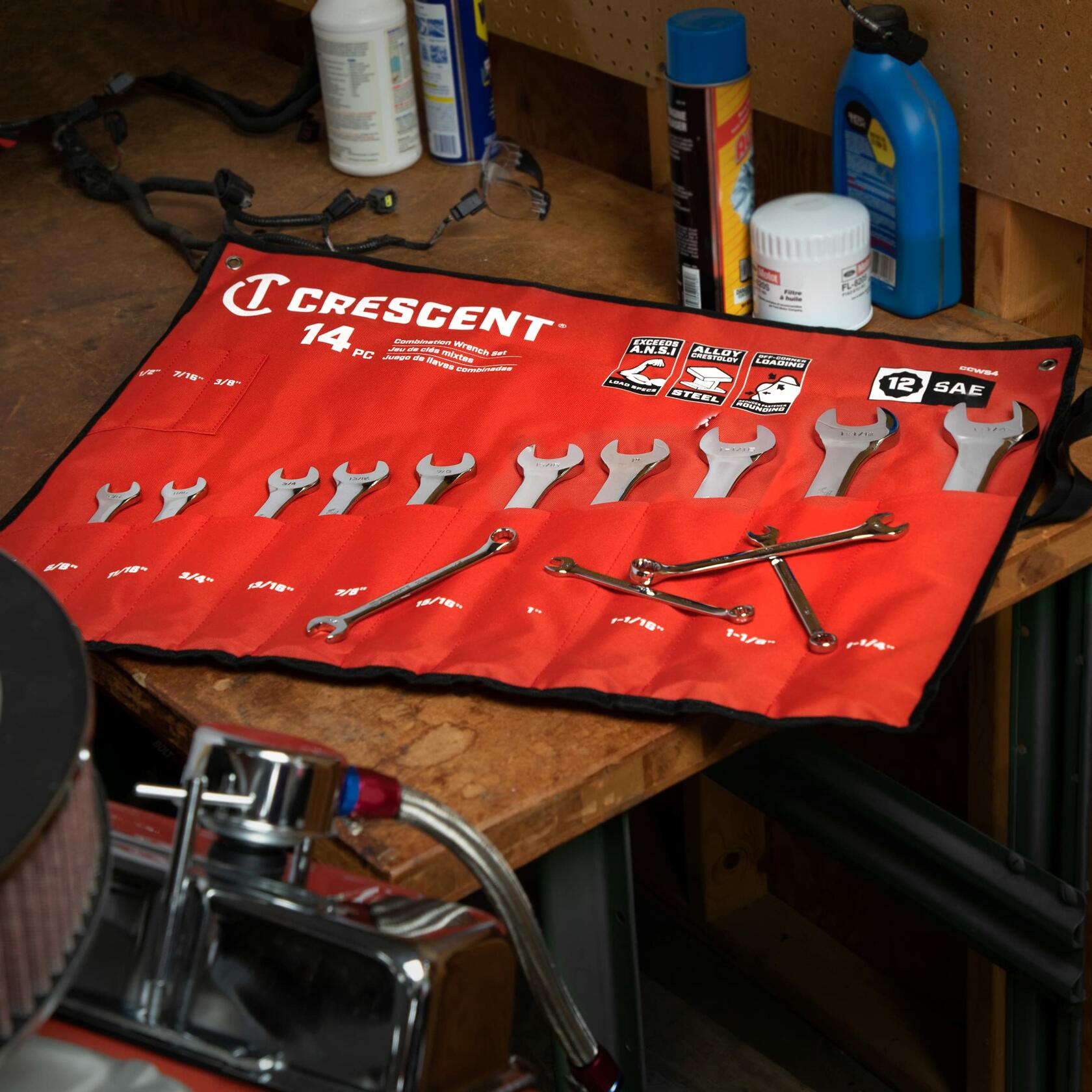 Crescent 14-piece SAE combination wrench set laid out on a workbench. Wrenches are stored in a red fabric tool roll labeled with sizes from 1/4" to 1-1/16". Surrounding items include motor oil, wipes, and workshop tools.