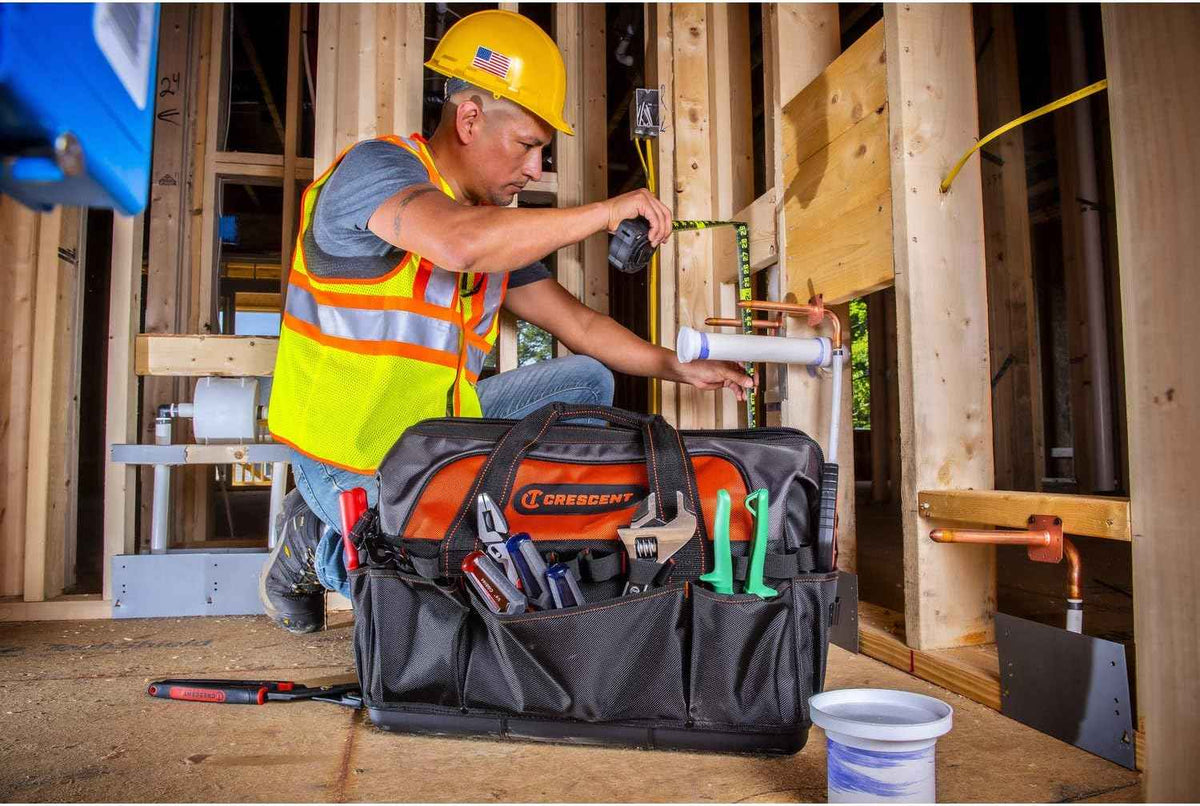 A contractor at a construction site measuring a piece of plumbing pipe next to the Crescent 20-inch tool bag, which holds various hand tools in its exterior pockets.