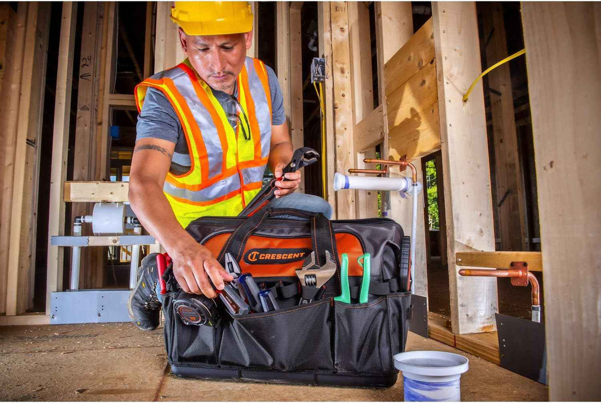 A contractor putting a tape measure into an exterior pocket of the Crescent 20-inch tool bag at a construction site, with various hand tools already organized in the remaining pockets.