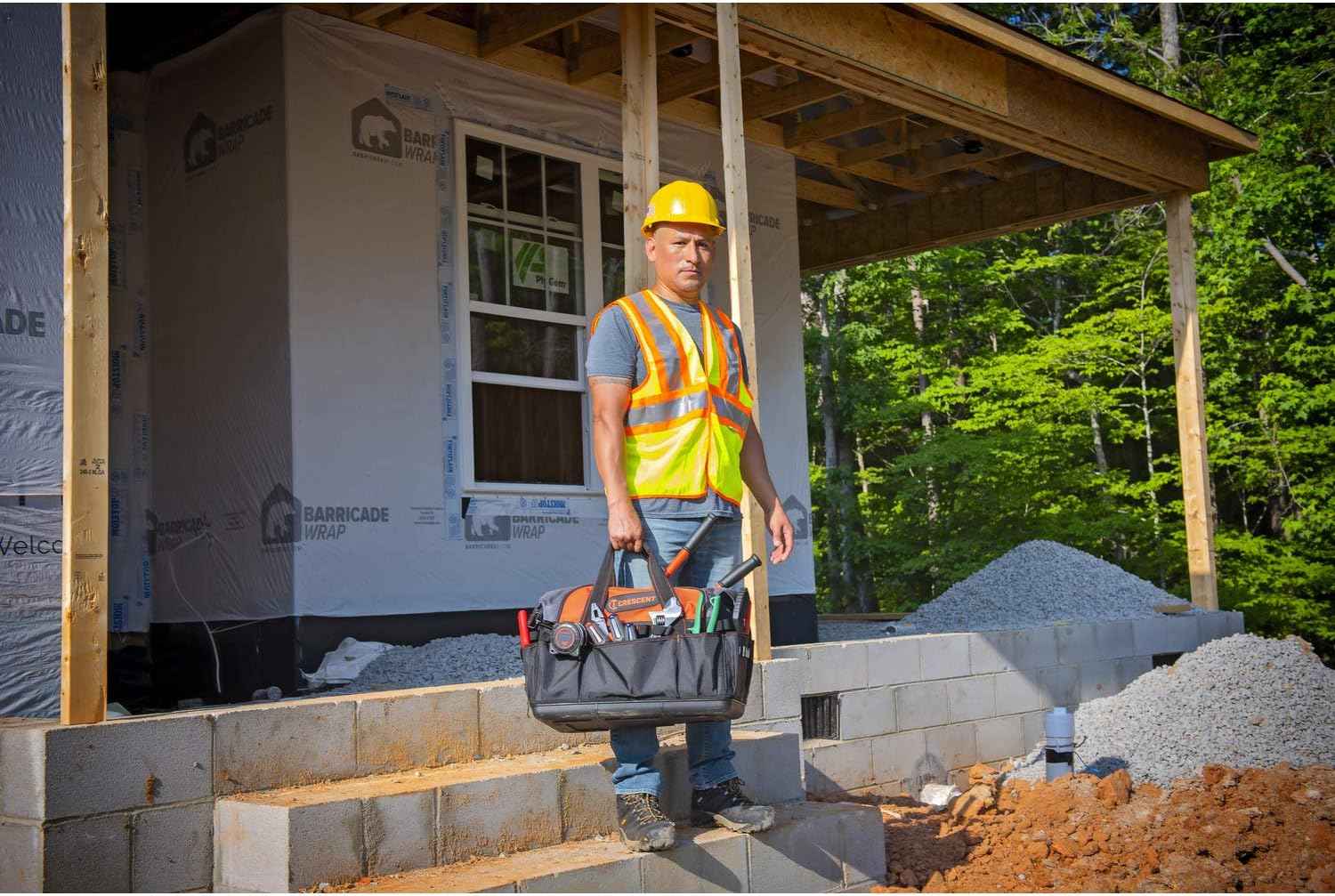 A contractor standing outdoors in front of a house under construction, holding the Crescent 20-inch tool bag by the padded handle, showcasing the bag's size and portability.