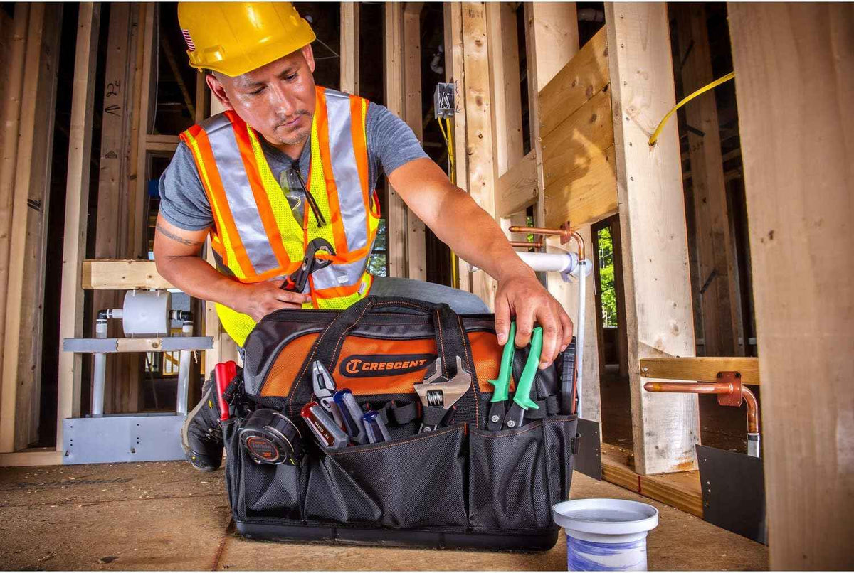 A contractor wearing a hard hat and safety vest kneeling at a construction site, placing a pair of pliers into one of the exterior pockets of the Crescent 20-inch tool bag, which is sitting on a wooden subfloor near copper piping.