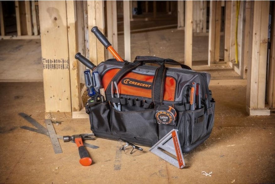 The Crescent 20-inch tool bag filled with tools sitting on a dusty wooden floor at a construction site, surrounded by a hammer, a square, safety glasses, and other carpentry tools.