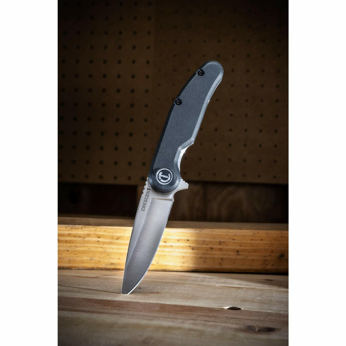 The open Crescent CPK325C pocket knife is shown resting with its blade tip stuck into a 2x4 on a wooden workbench.