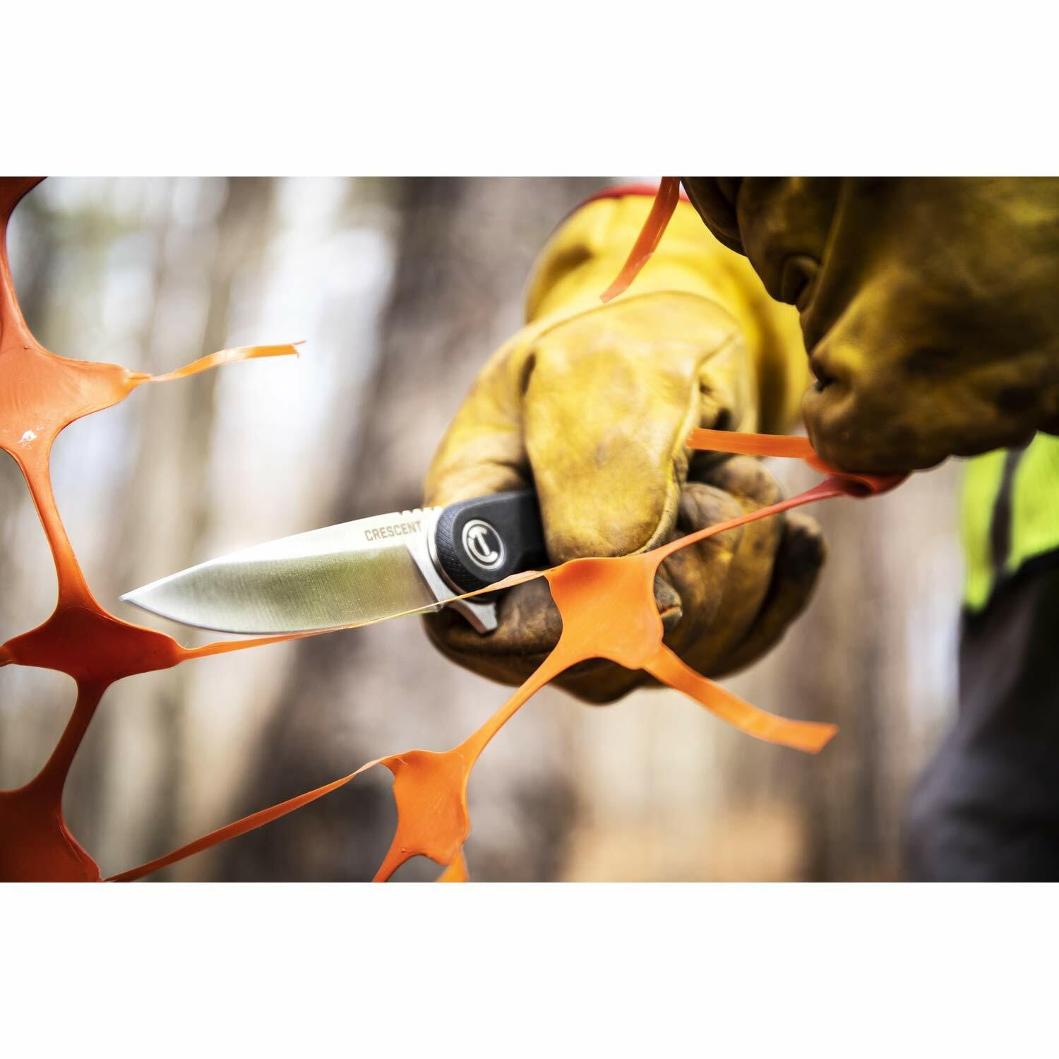 A gloved hand uses the Crescent CPK325C pocket knife to cut through a piece of orange plastic safety fencing.