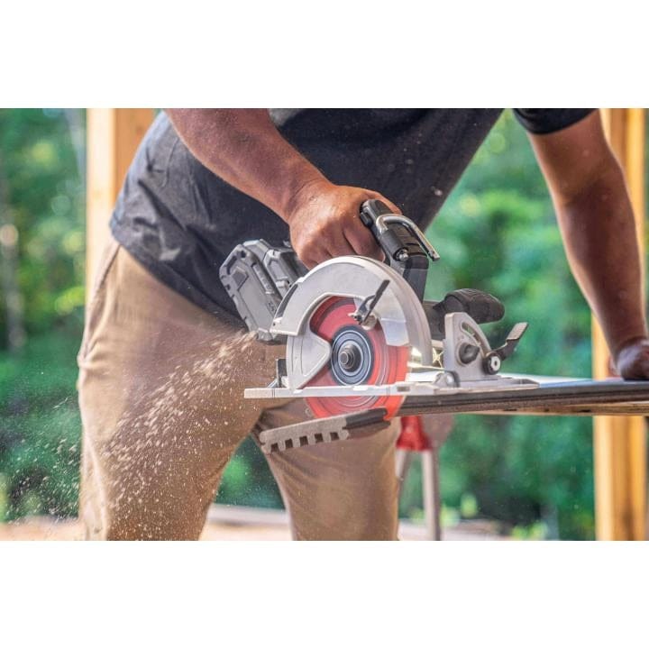 Action photo of a person using a handheld circular saw to cut wood outdoors. Sawdust is visible mid-cut, and the saw features a red and silver blade guard with a battery pack, suggesting cordless operation.