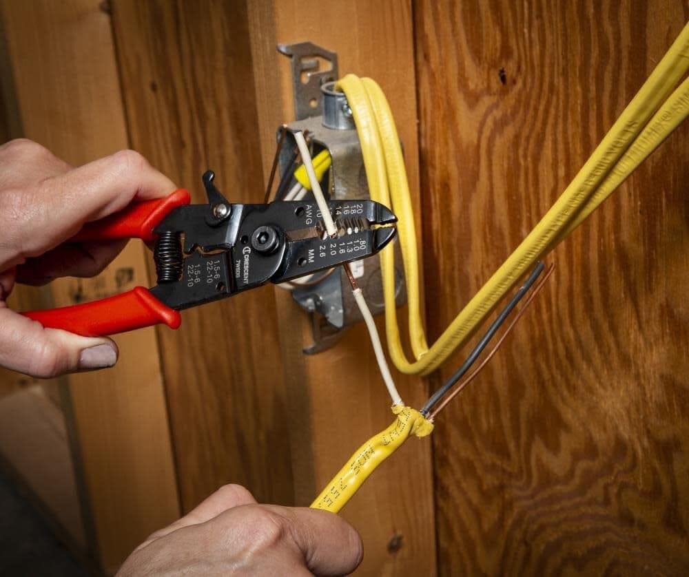 Application shot of an electrician using the Crescent 7WSDG pliers to strip insulation from a white electrical wire inside a metal junction box, demonstrating the tool's precision in a real-world environment.
