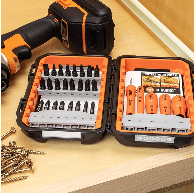 Open Crescent APEX fastening bit set displayed on a wooden surface. The left side contains black screwdriver bits labeled PH2, T25, and others. The right side features orange u-GUARD protected bits and nutsetters labeled with sizes like PH2, T25, 1/8", and 5/32". A promotional insert highlights the protective benefits of u-GUARD. A cordless drill and scattered screws are visible nearby.