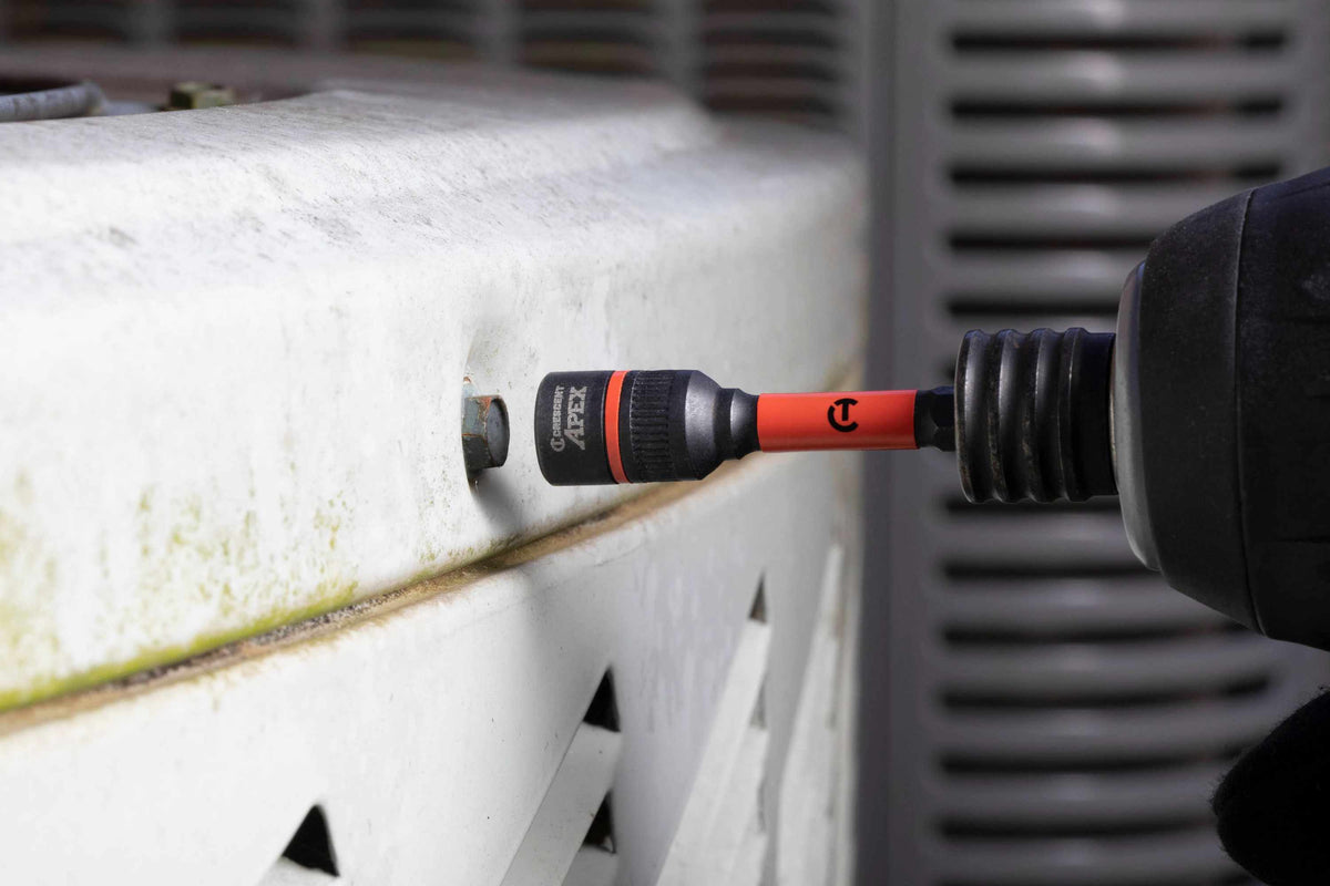 Action shot of the Crescent APEX 3/8-inch Bolt Biter attached to a cordless power impact driver, preparing to extract a rusted hex bolt from a white metal outdoor HVAC unit.