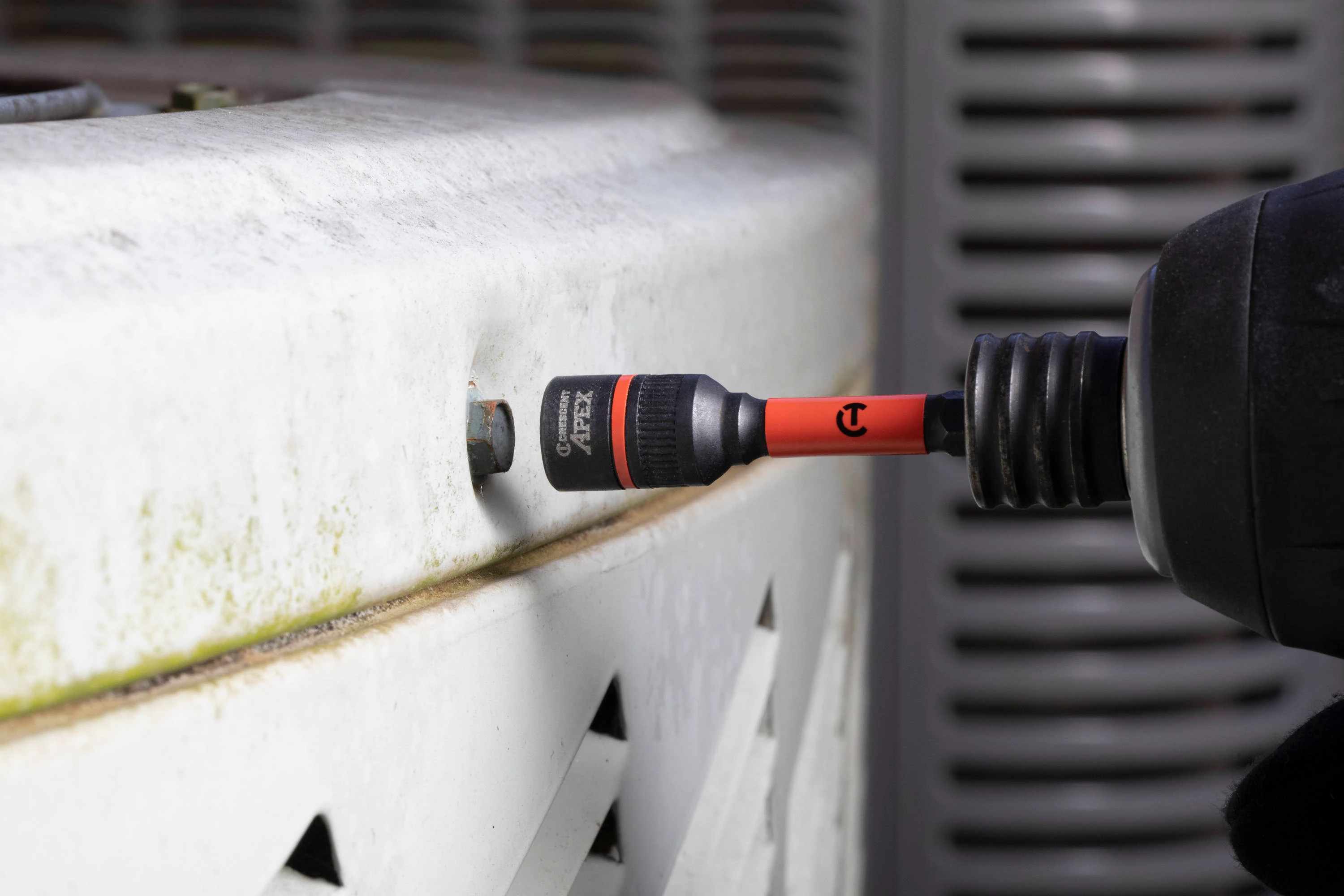 Action shot of the Crescent APEX 3/8-inch Bolt Biter attached to a cordless power impact driver, preparing to extract a rusted hex bolt from a white metal outdoor HVAC unit.