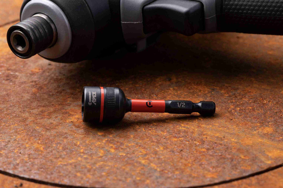 The Crescent APEX CAVBX2D32 1/2-inch Bolt Biter impact nut extractor resting on a rusted metal surface, showcasing its laser-etched 1/4-inch hex shank and orange impact zone, with a cordless impact driver blurred in the background.