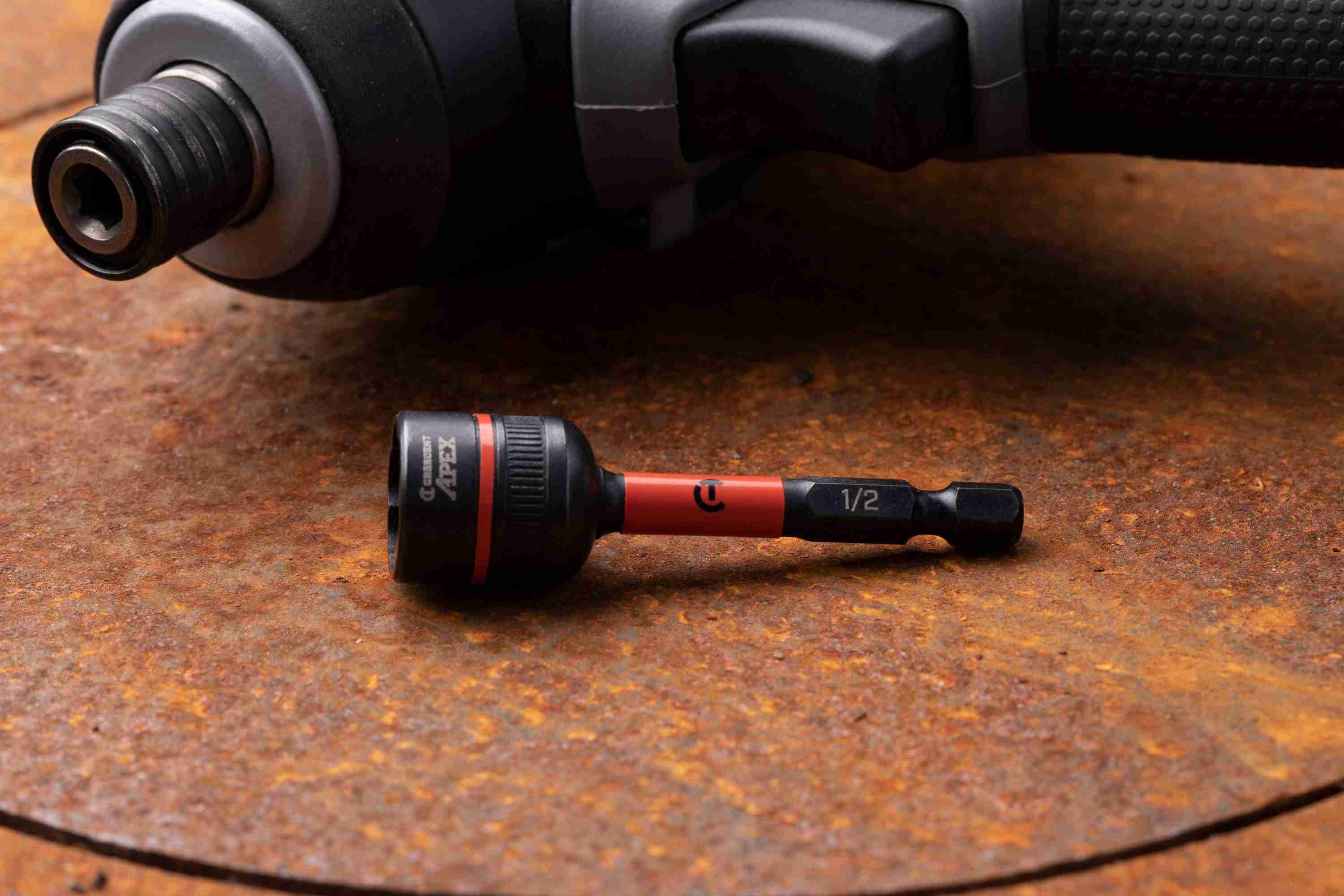 The Crescent APEX CAVBX2D32 1/2-inch Bolt Biter impact nut extractor resting on a rusted metal surface, showcasing its laser-etched 1/4-inch hex shank and orange impact zone, with a cordless impact driver blurred in the background.
