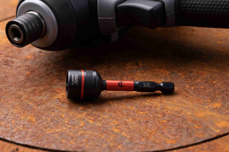 The Crescent APEX CAVBX2D32 1/2-inch Bolt Biter impact nut extractor resting on a rusted metal surface, showcasing its laser-etched 1/4-inch hex shank and orange impact zone, with a cordless impact driver blurred in the background.