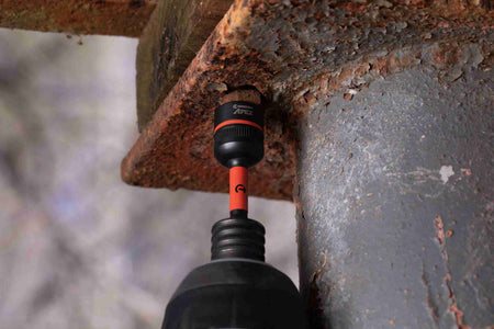 Action shot of the Crescent APEX 1/2-inch Bolt Biter attached to a cordless impact driver, about to engage a heavily rusted and corroded bolt securing a metal bracket to a weathered steel beam.