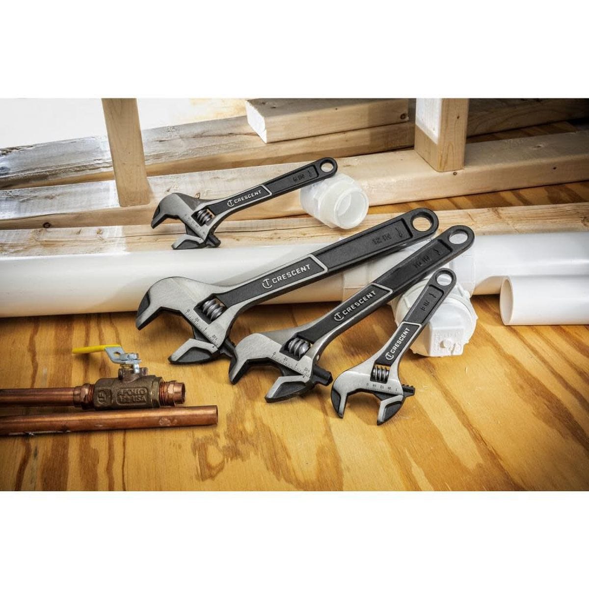 A jobsite lifestyle photograph displaying a family group of four various-sized black phosphate Crescent adjustable wrenches arranged neatly on a plywood deck next to copper pipes, a brass ball valve, and white PVC plumbing fittings.