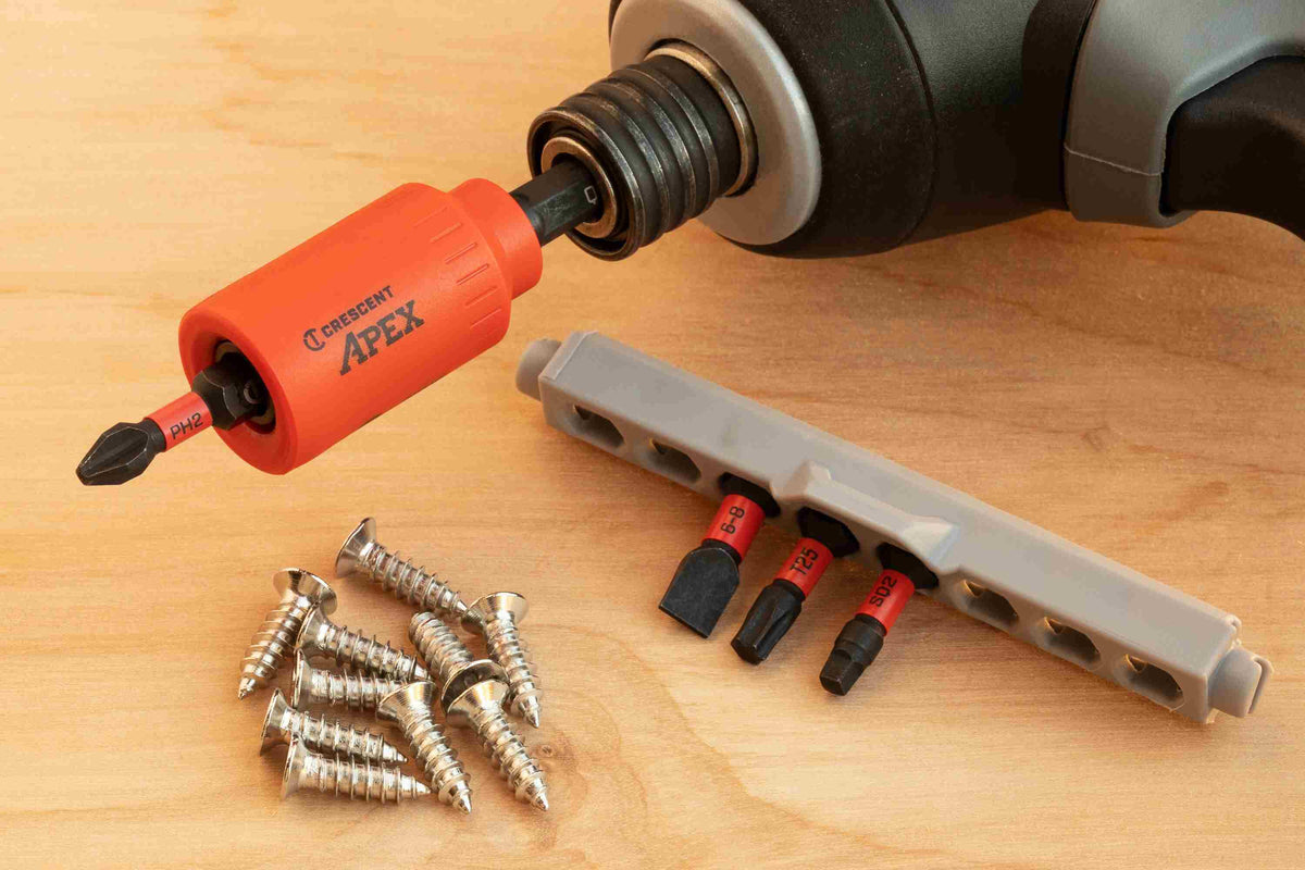 The Crescent u-GUARD non-marring bit holder attached to a cordless impact driver chuck, resting on a wooden workbench next to a pile of steel wood screws and the grey rail holding the remaining VORTEX impact power bits.