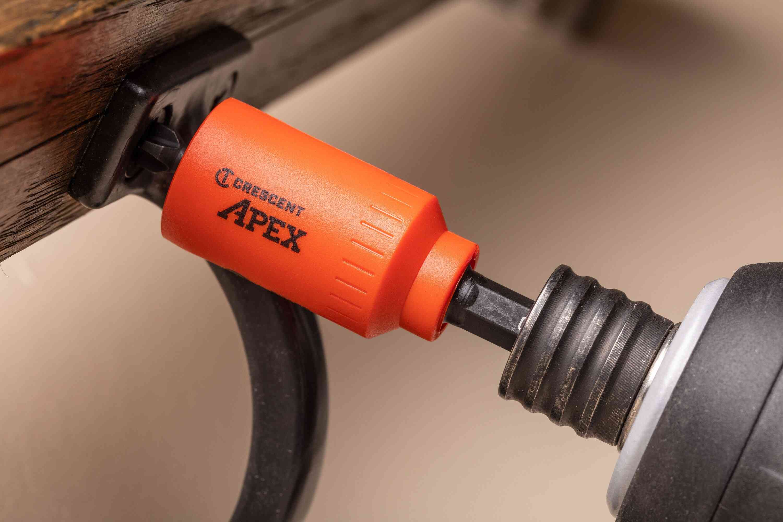 Close-up of an impact driver utilizing the Crescent u-GUARD quick-release bit holder to securely drive a fastener into a black metal bracket on rustic wood, showcasing the protective orange sleeve in tight clearance.