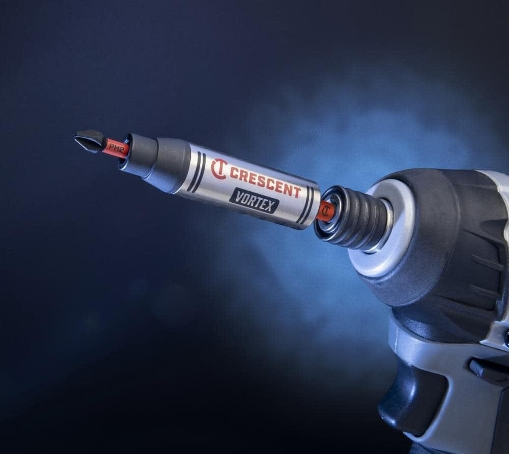 Dramatic studio shot of the Crescent VORTEX 1/4-inch hex shank bit holder attached to a power driver chuck, holding a PH2 Phillips insert bit against a dark blue background