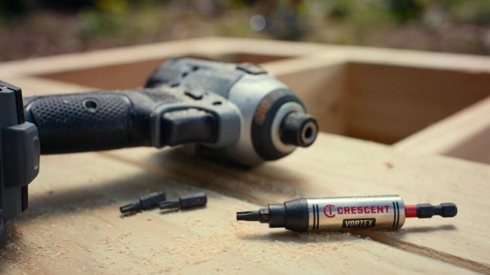 The Crescent CAVBH3 VORTEX impact bit holder and two extra hex bits resting on wooden framing lumber amidst sawdust, with a heavy-duty impact driver resting blurred in the background of a job site
