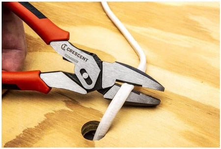 An action photo of the Crescent CCA20509 pivot action pliers being used to cut through a thick white electrical cable on a wooden workspace, demonstrating the tool's high-leverage cutting efficiency.