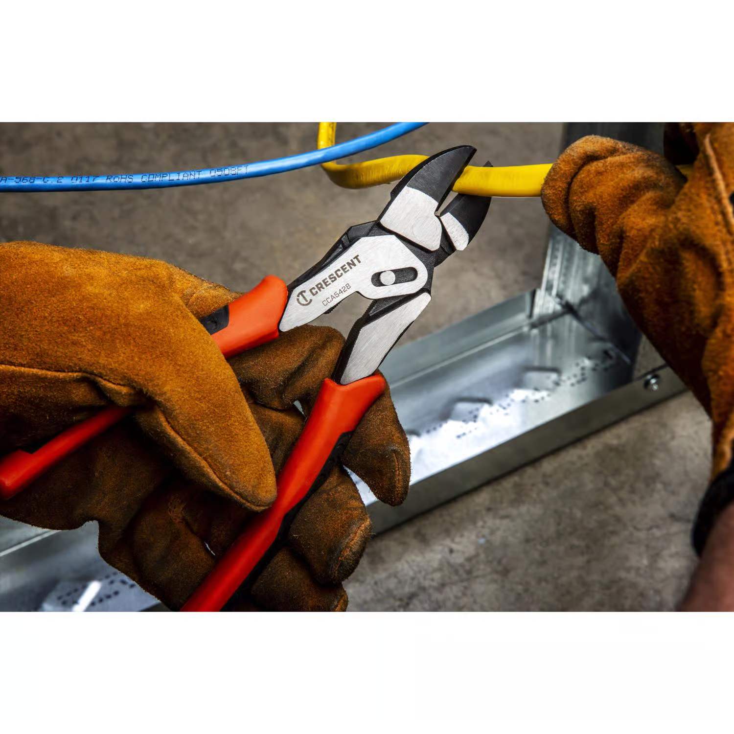 An action photo of a tradesperson using the Crescent CCA5428 pivot pro pliers to cut through a thick yellow electrical wire, demonstrating the tool's high-leverage compound action in a jobsite environment.