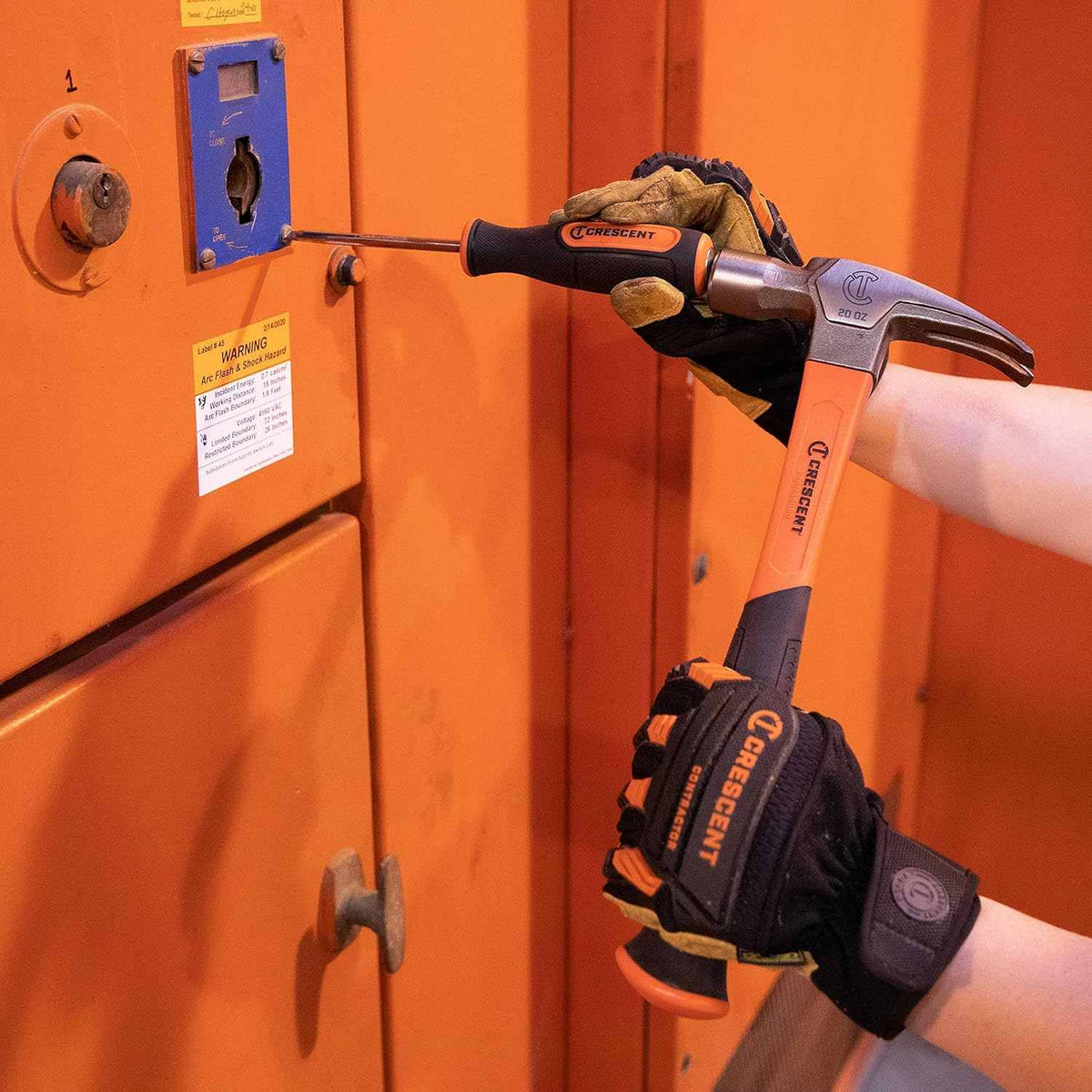 Close-up action shot of a worker's gloved hands striking the end cap of the Screw Biter screwdriver with a hammer to extract a screw from an orange metal panel.