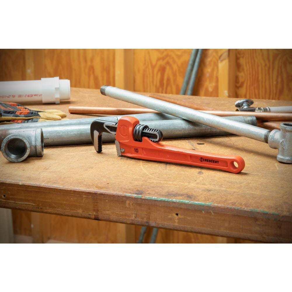 The Crescent CIPW10S cast iron pipe wrench resting on a wooden workbench among steel and copper pipes, illustrating its essential role in various plumbing and mechanical projects.