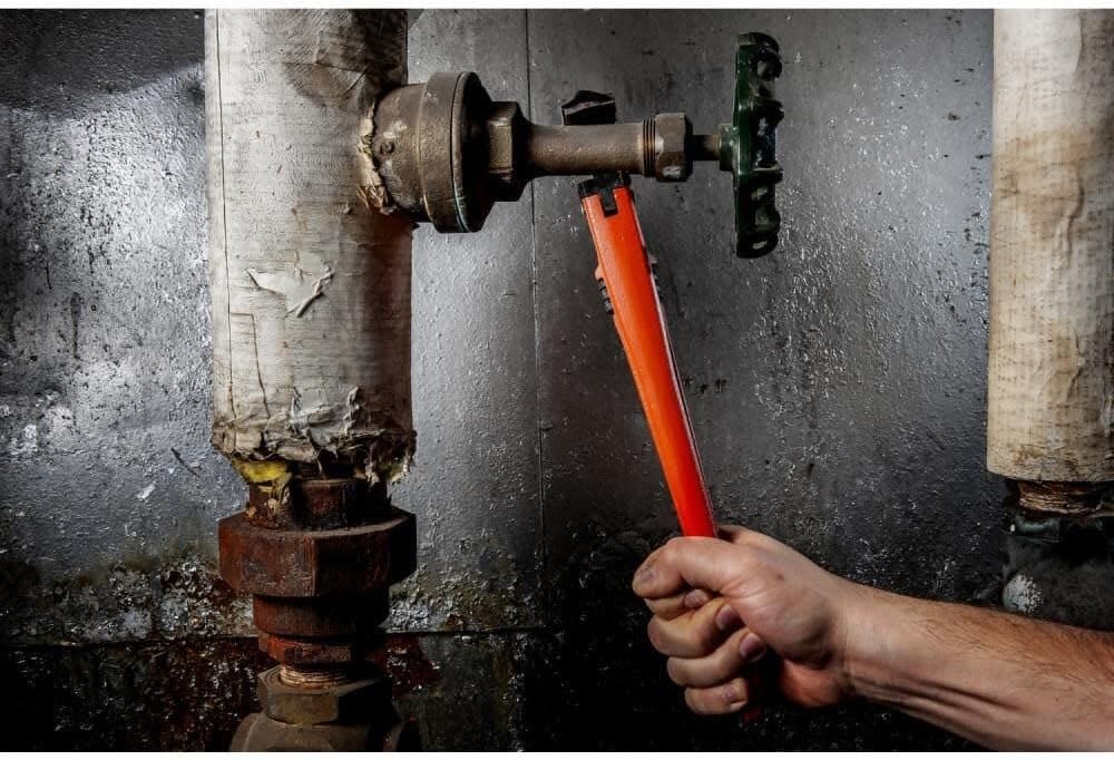 Vertical perspective of a plumber using the Crescent CIPW14 pipe wrench to adjust a valve fitting on an industrial piping system, illustrating the tool's reach and leverage.