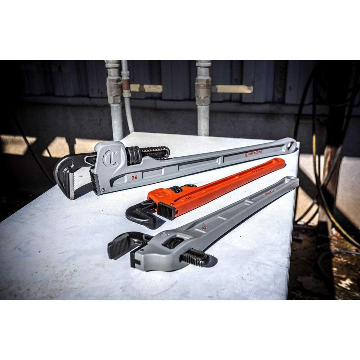 A real-world outdoor lifestyle layout showing a family of three different-sized heavy-duty Crescent plumbing wrenches arranged neatly on a metallic surface near electrical conduits and mechanical hookups.