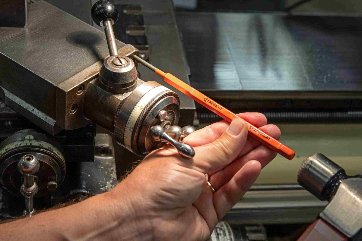 An action photograph of a professional using a Crescent pin punch on a metal lathe, demonstrating the tool's extended length and recessed control panel for accurate positioning in tight spaces.