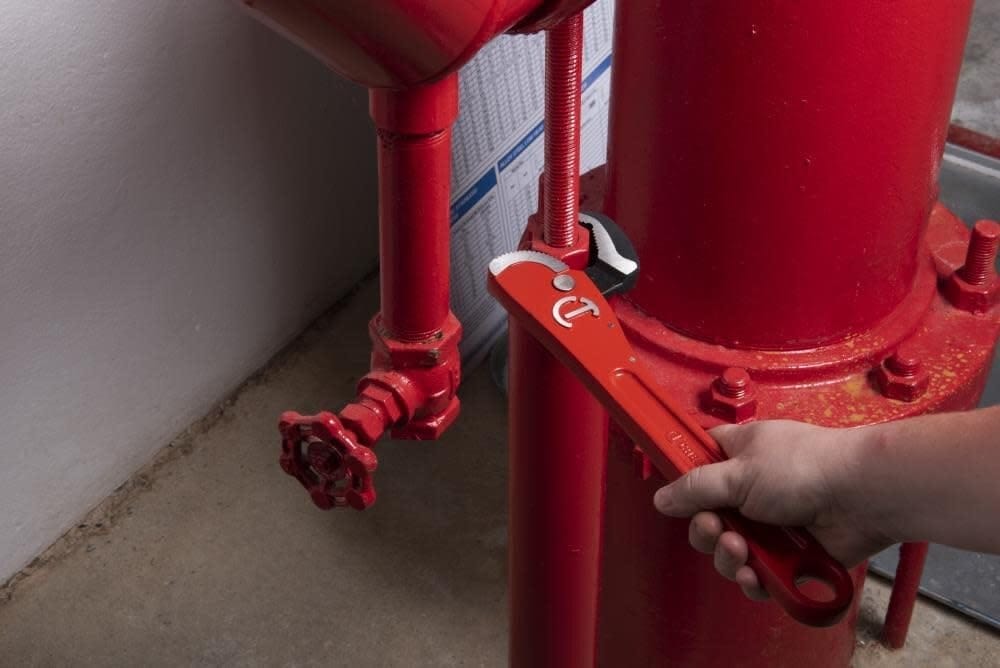 Side action view of the Crescent CPW12S wrench engaging a pipe fitting near a red industrial pump, highlighting the tool's effectiveness in reaching cramped utility spaces.