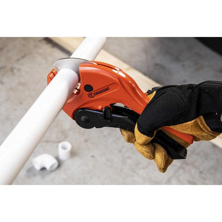 An action photograph of a professional using the Crescent CRPC118 ratcheting cutter to slice through a white PVC pipe, demonstrating the tool's one-handed operation and precision performance in a plumbing installation setting.