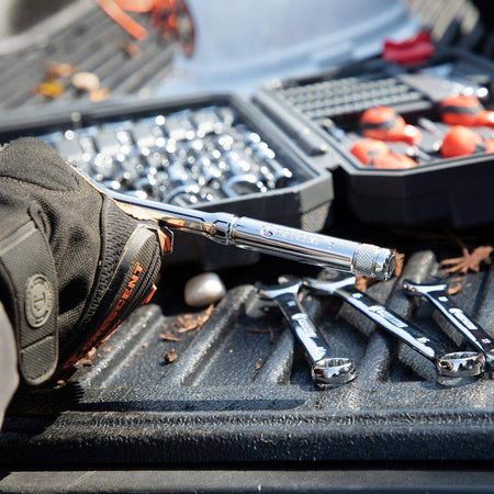 An action-oriented perspective of a gloved hand holding a Crescent ratchet over a truck bed, with various wrenches and the open tool case in the background, showcasing real-world jobsite utility.