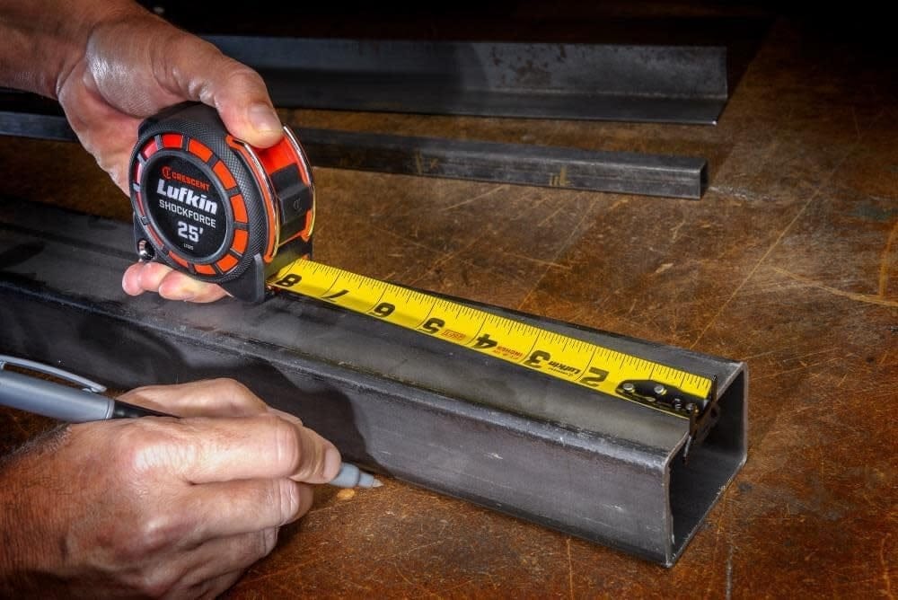 Action shot of a trade professional using the Crescent Shockforce tape measure and a marker to perform layout work on a metal square tube in a workshop setting.