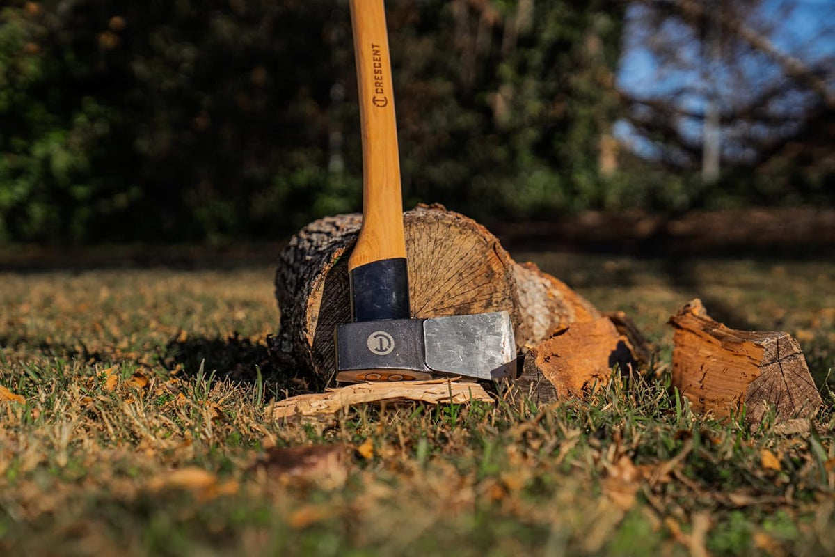 A functional application shot of the Crescent wood chopping axe embedded in a log, demonstrating its industrial cutting power and durable construction in an outdoor environment.