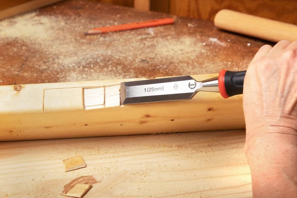 An action shot of a craftsman using a Crescent 1-inch wood chisel to carve a precision notch into a wooden beam. The image demonstrates the tool's effectiveness for jobsite joinery, showcasing the sharp carbon steel blade as it removes wood shavings on a workbench.