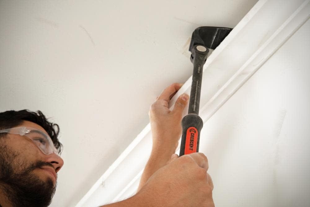 Action shot of a person using the Crescent adjustable pry bar to carefully pry white crown molding away from a ceiling corner without damaging the surface.
